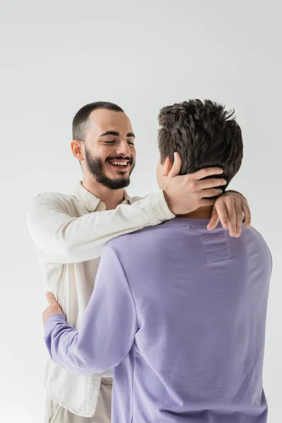 Hombre homosexual barbudo positivo con los ojos cerrados en camisa casual tocando el cuello y abrazando novio morena mientras está de pie aislado en gris - foto de stock