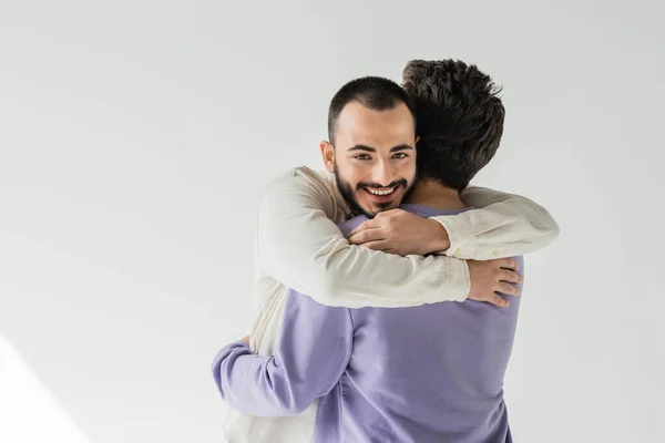 Alegre e barbudo gay homem no casual roupas abraçando jovem morena namorado e olhando para câmera no cinza fundo com luz solar — Fotografia de Stock