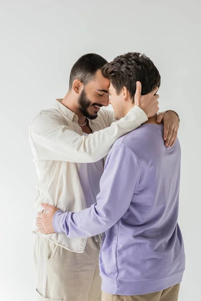 Smiling and bearded gay man in casual beige clothes closing eyes while touching neck and hugging brunette boyfriend isolated on grey — Stock Photo
