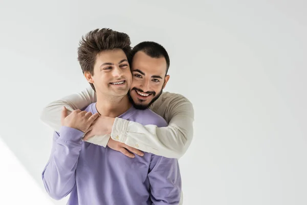 Веселий і бородатий гомосексуальний чоловік приймає молодого брюнетки-партнера в брекети і фіолетовий светр, дивлячись на камеру на сірому фоні з сонячним світлом — стокове фото