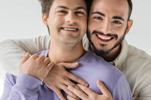 Retrato de jovem e alegre casal do mesmo sexo em roupas casuais abraçando e tocando as mãos enquanto olha para a câmera isolada no cinza — Fotografia de Stock