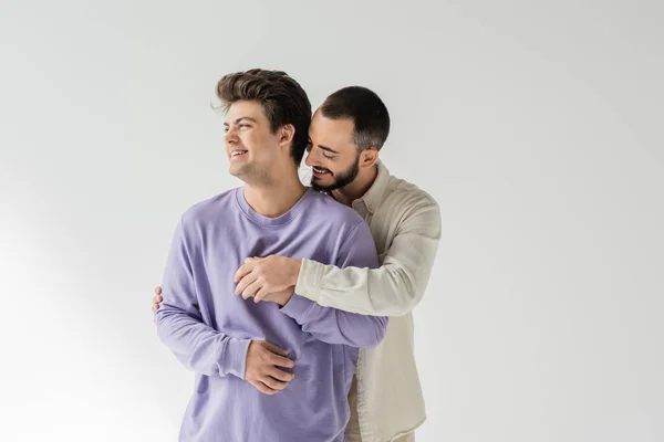 Homme homosexuel barbu en vêtements décontractés étreignant et tenant la main de jeune partenaire brune en bretelles et sweat-shirt violet tout en étant isolé sur gris — Photo de stock