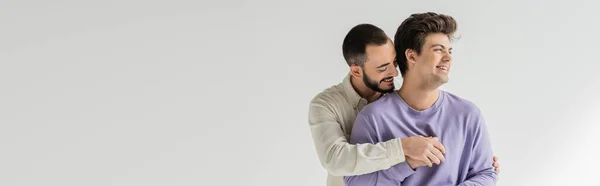 Young bearded homosexual man hugging carefree brunette boyfriend in braces and sweatshirt looking away while standing isolated on grey, banner — Stock Photo