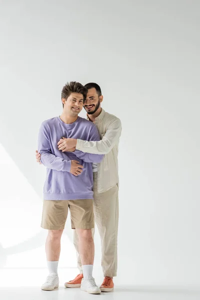 Full length of positive same sex couple in casual clothes holding hands and looking at camera while standing on grey background with sunlight — Stock Photo