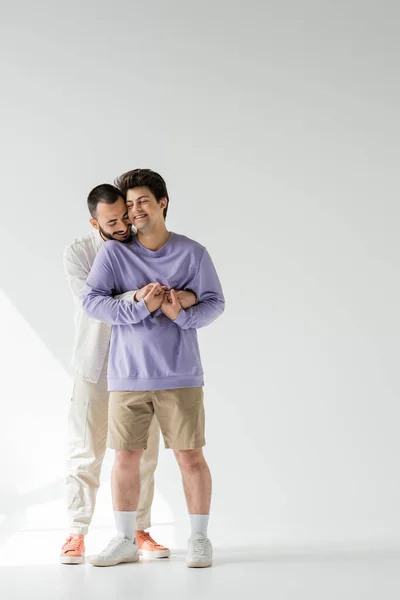 Hombre homosexual sonriente y barbudo con ropa casual cogido de la mano de un joven novio con los ojos cerrados mientras está de pie sobre un fondo gris con luz solar - foto de stock