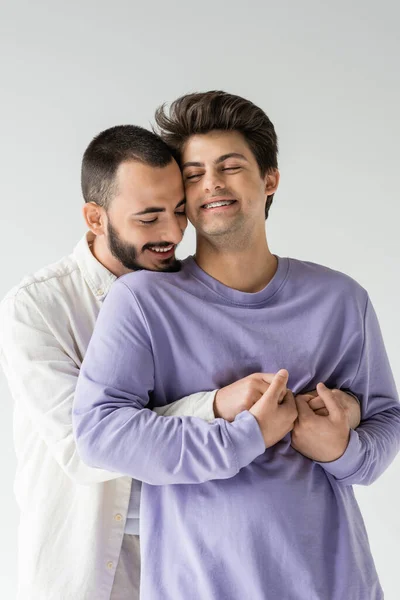 Überglücklicher und bärtiger schwuler Mann, der Hände hält und seinen brünetten Freund mit Zahnspange und geschlossenen Augen umarmt, während er isoliert auf grau steht — Stockfoto