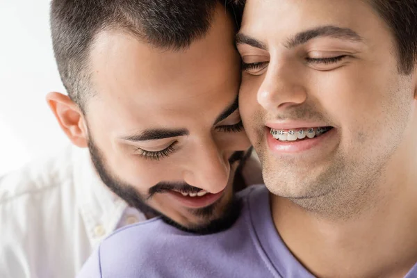 Close up vista di sorridente e barbuto gay uomo in piedi vicino a giovane partner con gli occhi chiusi e bretelle sui denti isolati su grigio — Foto stock