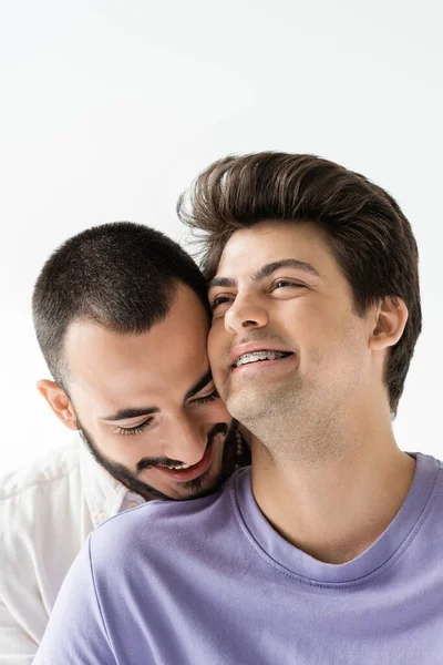 Retrato de morena y hombre gay positivo con tirantes en los dientes mirando hacia otro lado mientras está de pie cerca de novio barbudo aislado en gris - foto de stock