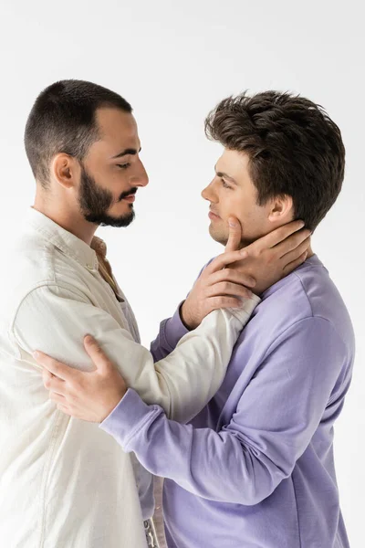 Вид сбоку на нежного гомосексуалиста в повседневной одежде, трогающей шею и смотрящего на молодого парня брюнетки, стоящего в изоляции на серой — стоковое фото