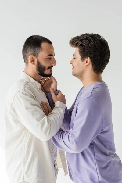 Vista lateral de homem homossexual morena exultante com olhos fechados tocando bochecha de namorado barbudo em roupas casuais isolado em cinza — Fotografia de Stock