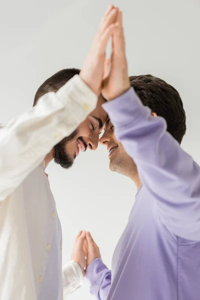 Low angle view of young and smiling homosexual couple in casual clothes closing eyes while holding hands and standing together isolated on grey — Stock Photo