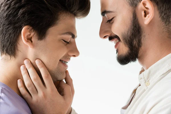 Vista lateral del hombre gay barbudo positivo en camisa casual tocando la cara de la joven pareja con tirantes en los dientes mientras están de pie juntos aislados en gris - foto de stock