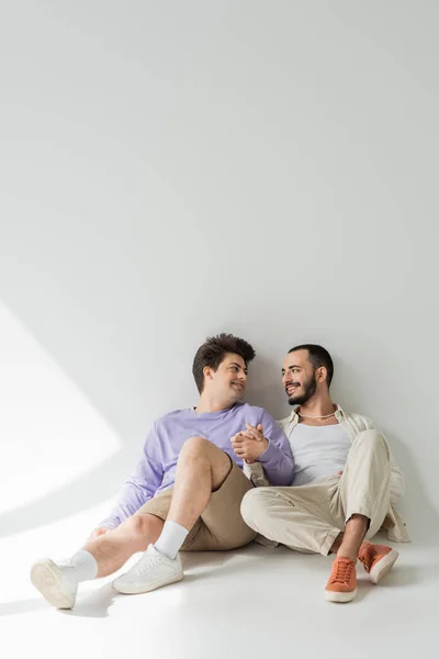 Full length of smiling homosexual couple in casual clothes holding hands and looking at each other while sitting together on grey background with sunlight — Stock Photo