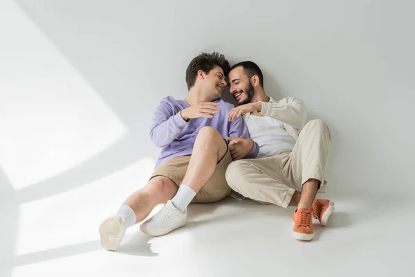 Full length of cheerful same sex partners with closed eyes holding hands while talking and sitting together on grey background with sunlight — Stock Photo
