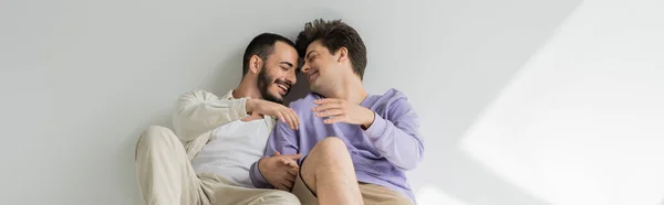 Riendo pareja homosexual con los ojos cerrados tomados de la mano y conversando mientras están sentados juntos sobre un fondo gris con luz solar, pancarta - foto de stock