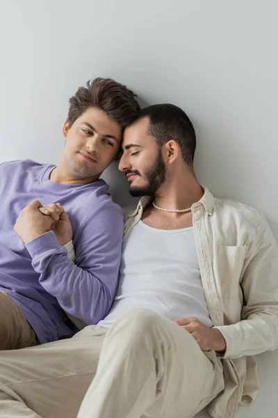 Young homosexual man in sweatshirt holding hand and looking at bearded boyfriend with closed eyes while sitting on grey background — Stock Photo