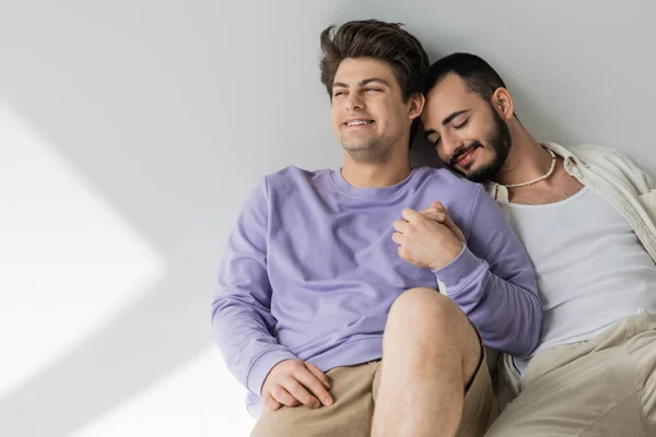 Sorrindo jovem gay homem em roupas casuais segurando mão de namorado barbudo com olhos fechados enquanto sentados juntos no fundo cinza com luz solar — Fotografia de Stock