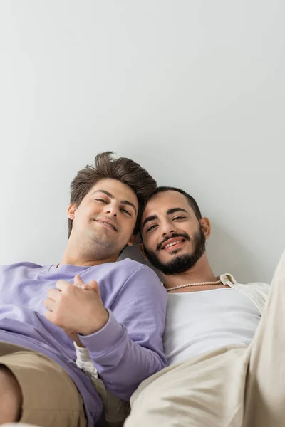 Позитивные и молодые однополые партнеры в повседневной одежде держатся за руки и смотрят в камеру, лежа и расслабляясь вместе на сером фоне — стоковое фото