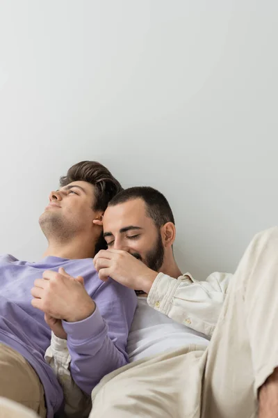 Joven homosexual tocando hombro y cogido de la mano de novio morena en ropa casual mientras se sientan juntos sobre fondo gris - foto de stock