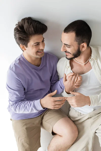 High angle view of young and cheerful homosexual couple in casual clothes holding hands while talking and sitting together on grey background — Stock Photo