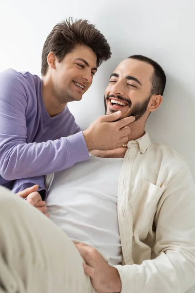 Sorridente gay uomo con bretelle su denti toccare faccia di barbuto bruna fidanzato con chiuso gli occhi e tenendo le mani mentre seduta insieme su sfondo grigio — Foto stock