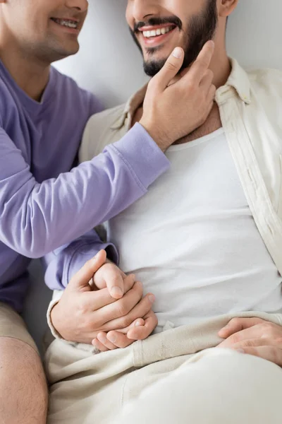 Cropped view of blurred and smiling man in braces sitting in casual clothes and holding hands with boyfriend while sitting together on grey background — Stock Photo