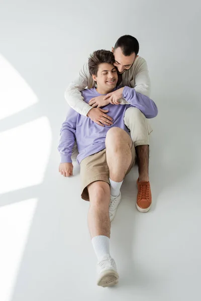 High angle view of brunette gay man in casual clothes embracing cheerful partner in braces with closed eyes while sitting together on grey background — Stock Photo