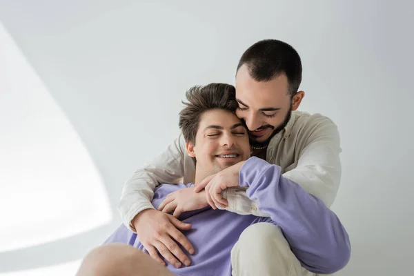Молодой брюнетка гей обнимает беззаботного парня с закрытыми глазами, в брекетах и в фиолетовой толстовке, сидя вместе на сером фоне с солнечным светом — стоковое фото