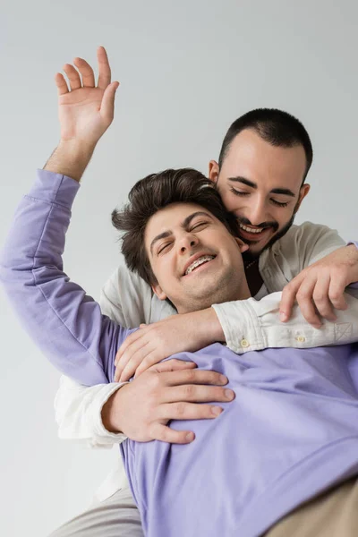 Positiver und bärtiger homosexueller Mann in lässiger Kleidung umarmt brünetten Partner mit geschlossenen Augen und Zahnspange, während er isoliert auf grau sitzt — Stockfoto