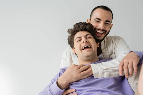 Бородатый гомосексуалист в рубашке обнимает и трогает руку смеющегося бойфренда в брекетах и фиолетовой толстовке, сидя изолированно на сером — стоковое фото