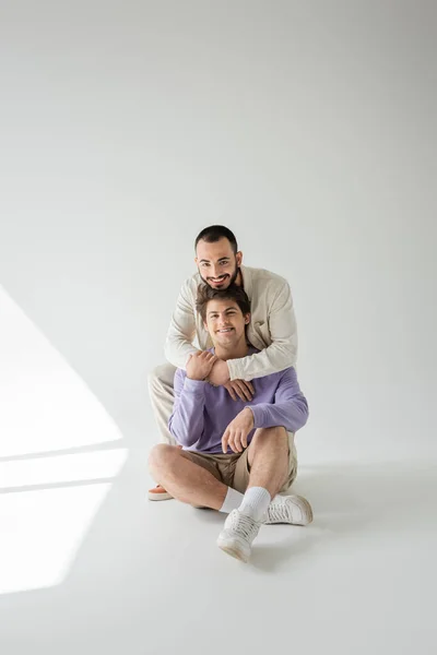 Joven pareja gay sonriente en ropa casual abrazando y mirando a la cámara mientras están sentados juntos sobre un fondo gris con luz solar - foto de stock