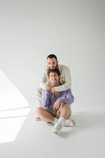 Young smiling gay couple in casual clothes hugging and looking at camera while sitting together on grey background with sunlight — Stock Photo
