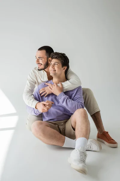 Junges und unbeschwertes gleichgeschlechtliches Paar, das sich umarmt und wegsieht, während es auf grauem Hintergrund mit Sonnenlicht und Schatten sitzt und entspannt — Stockfoto