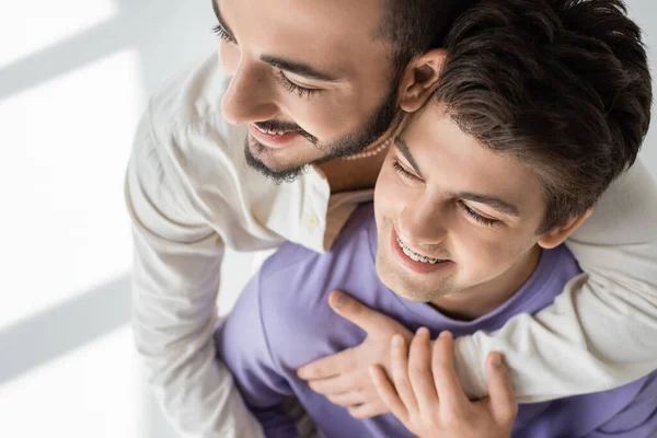 Vista aérea del hombre homosexual positivo y barbudo abrazando al joven novio en sudadera púrpura y tirantes sobre fondo gris con luz solar - foto de stock