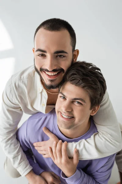Retrato de jóvenes novios homosexuales alegres con ropa casual abrazando y tocando las manos mientras miran a la cámara sobre un fondo gris con luz solar - foto de stock