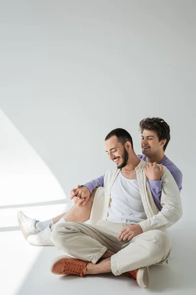 Joven gay hombre sonriendo mientras toma de la mano y abrazando barbudo novio en casual camisa mientras sentado en gris fondo con sol - foto de stock
