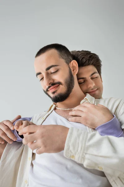 Portrait of smiling and bearded homosexual man in casual clothes and pearl necklace touching hand of brunette boyfriend closing eyes isolated on grey — Stock Photo