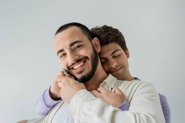 Portrait of positive and bearded gay man holding hand of young brunette boyfriend near face and looking at camera isolated on grey — Stock Photo