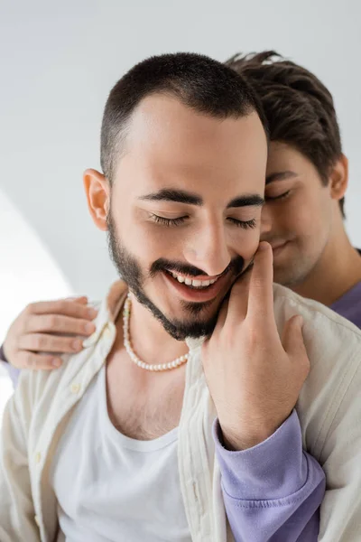Размытый и брюнетка гей мужчина обнимает и трогает щеку сверх радостного и бородатого партнера с закрытыми глазами на сером фоне — стоковое фото