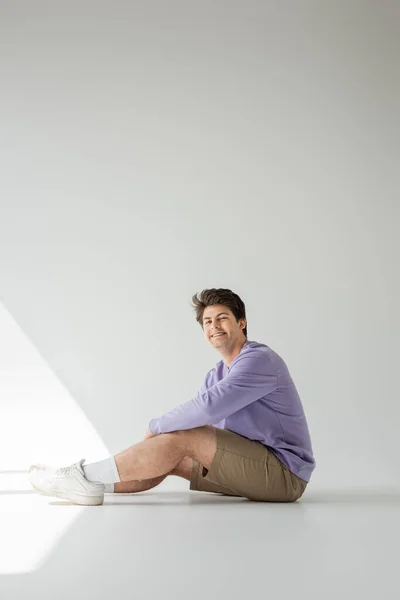 Longitud completa del hombre homosexual sonriente con tirantes, pantalones cortos beige y sudadera púrpura mirando a la cámara mientras está sentado sobre un fondo gris con luz solar - foto de stock
