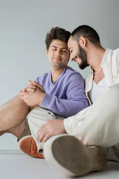 Joven y positiva pareja del mismo sexo con los ojos cerrados en ropa casual tomados de la mano mientras están sentados y descansando juntos sobre un fondo gris - foto de stock