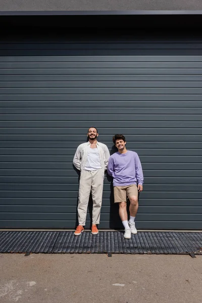 Full length of cheerful same sex couple in casual clothes looking at camera while standing near wall of grey building on urban street — Stock Photo
