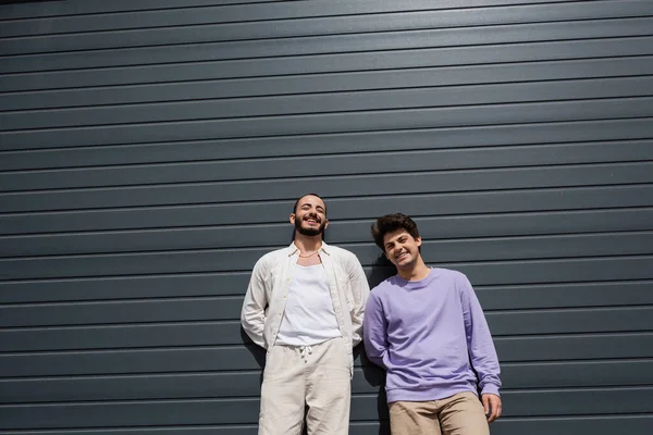 Joven y positiva pareja homosexual en traje casual mirando a la cámara mientras están de pie juntos cerca de la pared del edificio en la calle urbana durante el día - foto de stock