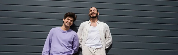 Alegre y joven pareja homosexual en ropa casual sonriendo a la cámara mientras está de pie cerca de la pared gris del edificio al aire libre durante el día, pancarta - foto de stock