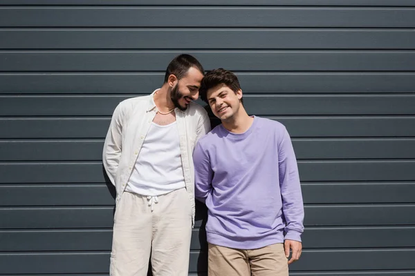 Lächelnder und bärtiger schwuler Mann in lässiger Kleidung, der neben einem jungen Freund in Hosenträgern steht und tagsüber in Kamera und Wand eines grauen Gebäudes im Freien blickt — Stockfoto