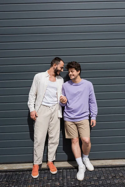 Full length of positive and young same sex couple in casual clothes holding hands while standing together near grey building on urban street — Stock Photo