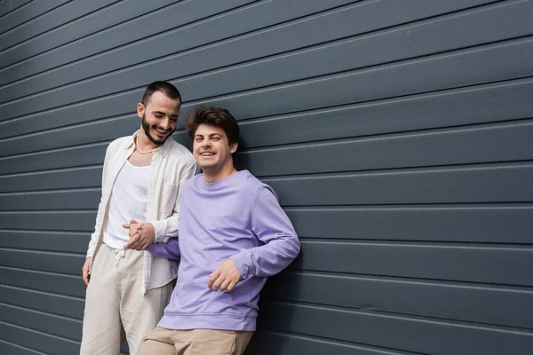 Cheerful and young gay man in sweatshirt and braces holding hand of bearded boyfriend while standing together near wall of building on urban street — Stock Photo