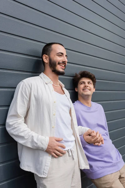 Homosexual bearded man smiling and looking away while holding hand of young boyfriend in braces near wall of building outdoors at daytime — Stock Photo