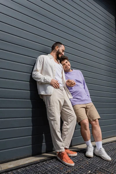 Full length of cheerful same sex couple in casual clothes holding hands while standing together near grey building outdoors at daytime — Stock Photo