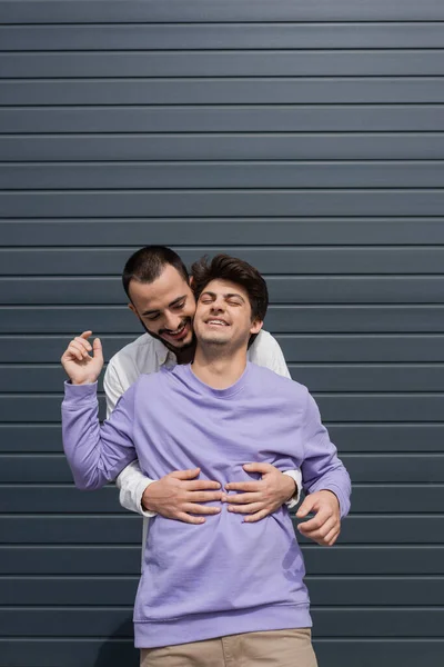 Homem homossexual alegre e barbudo abraçando parceiro morena no aparelho rindo enquanto estava perto de construção na rua urbana durante o dia — Fotografia de Stock